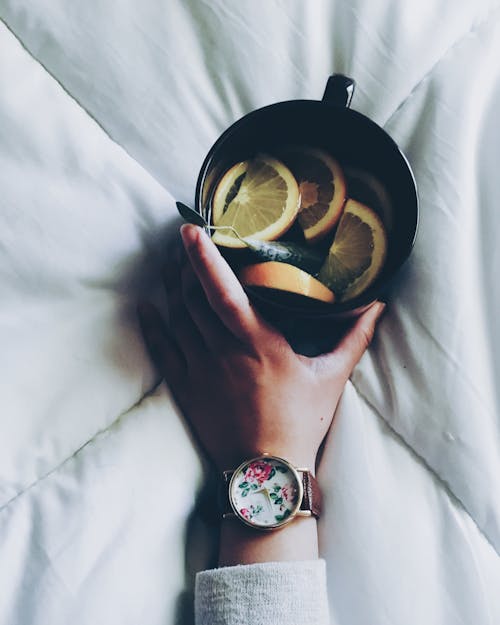 Donna Che Tiene La Tazza In Ceramica Nera Con Fette Di Limone Sulla Sommità Del Piumino Del Letto Bianco