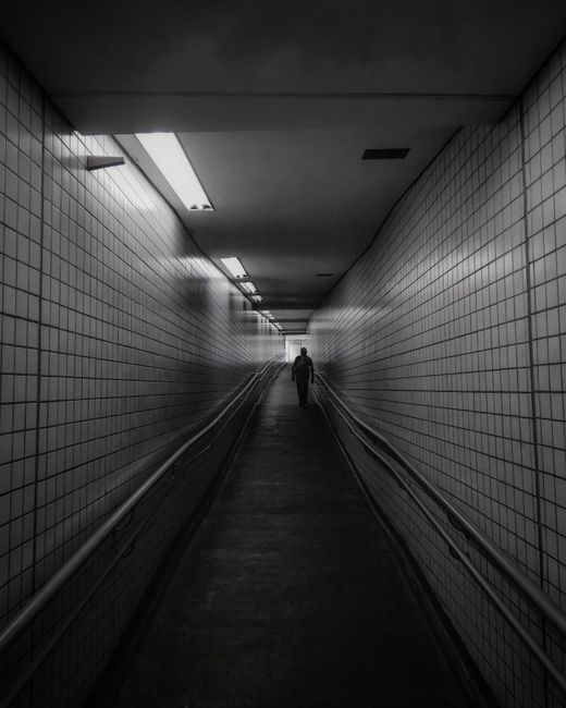 A solitary figure walks through a dimly lit underground tiled tunnel.
