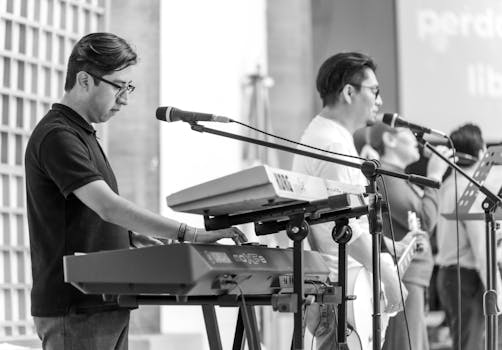 Musicians performing live with keyboard and guitar in a black and white concert setting.