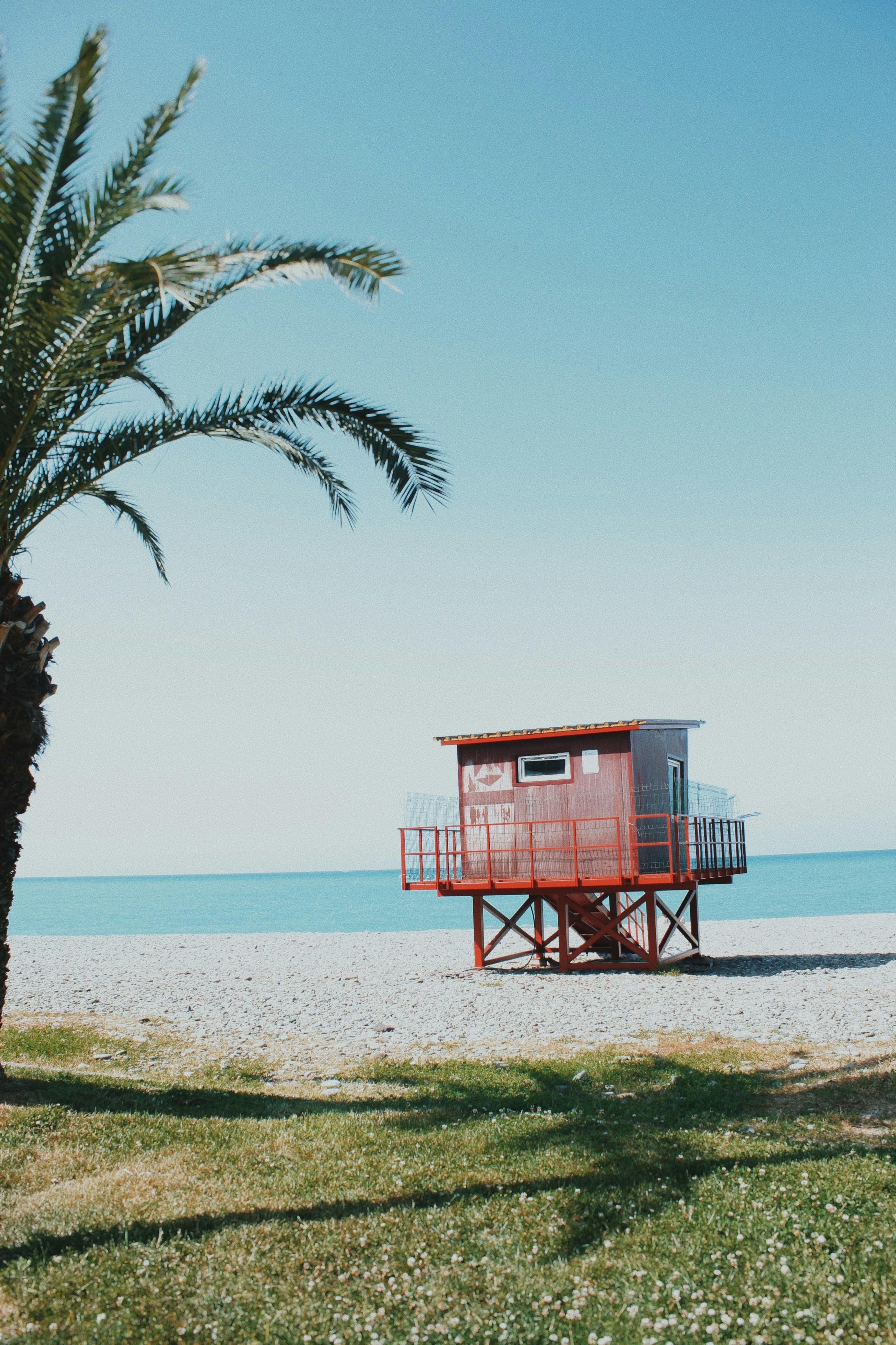 A tranquil beach scene featuring a red lifeguard hut and palm tree under a clear blue sky.