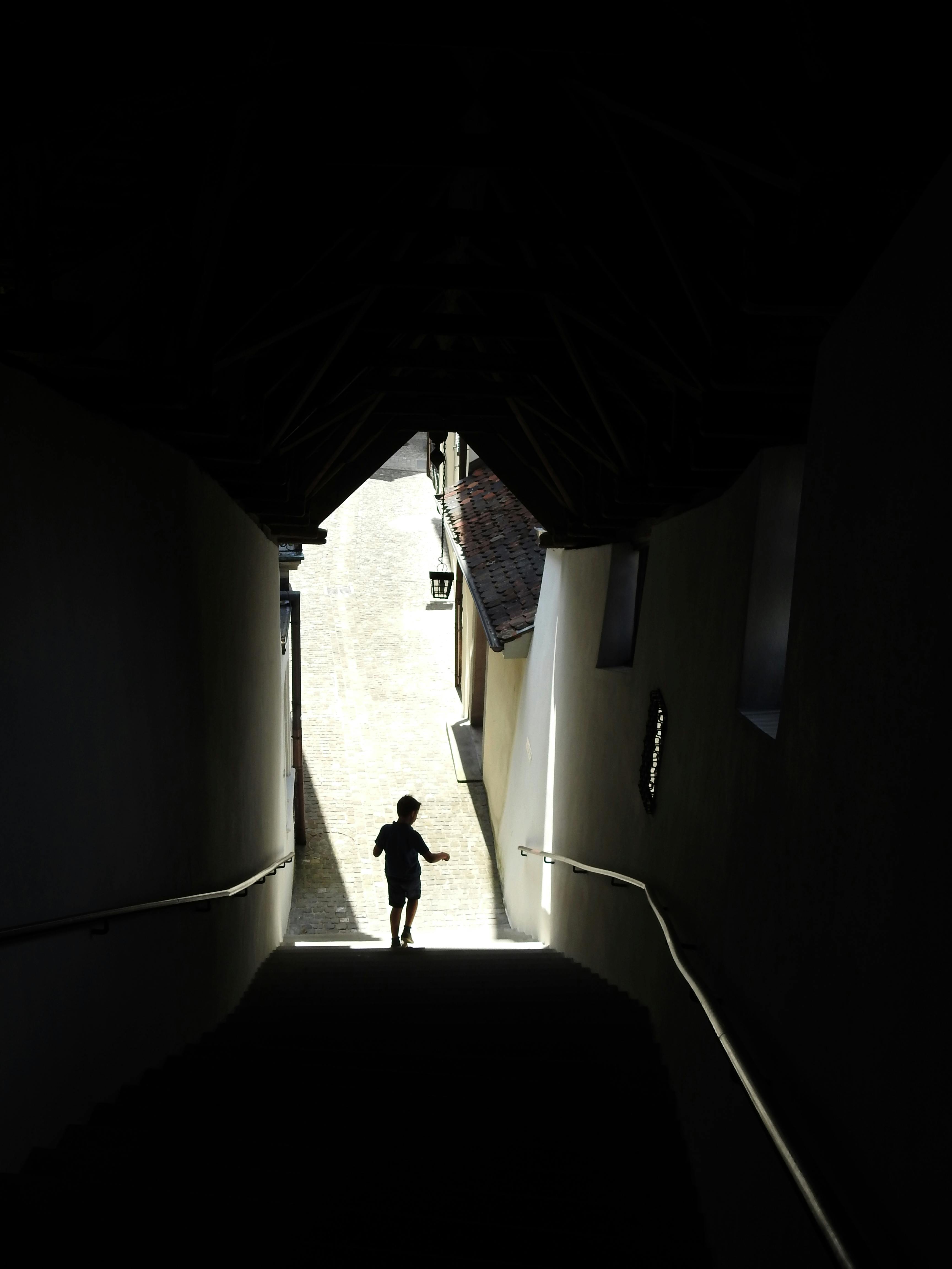A child in silhouette walks down a dimly lit urban stairway into bright daylight.
