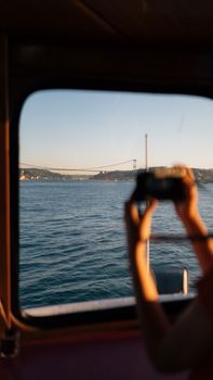 View of the Bosphorus Bridge from a ferry window in Istanbul, Turkey, with a person taking a photo.