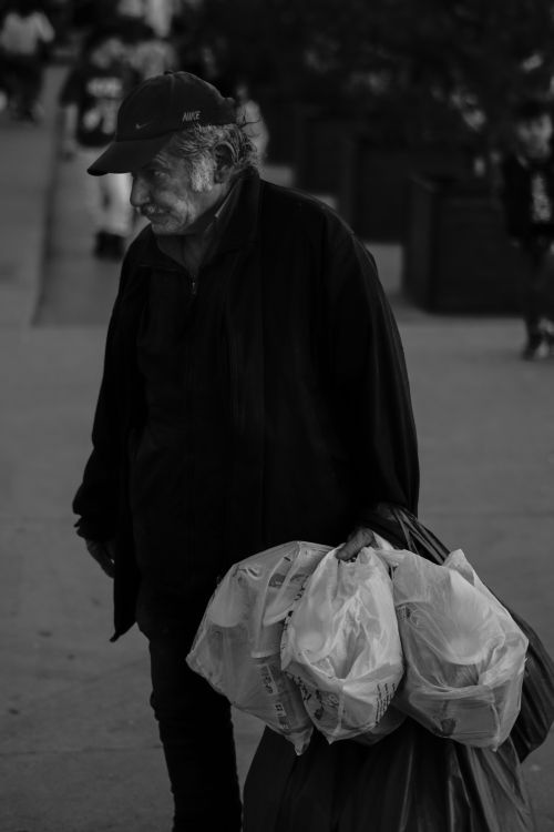 Free A black and white photo of an elderly man carrying plastic bags on an urban street. Stock Photo