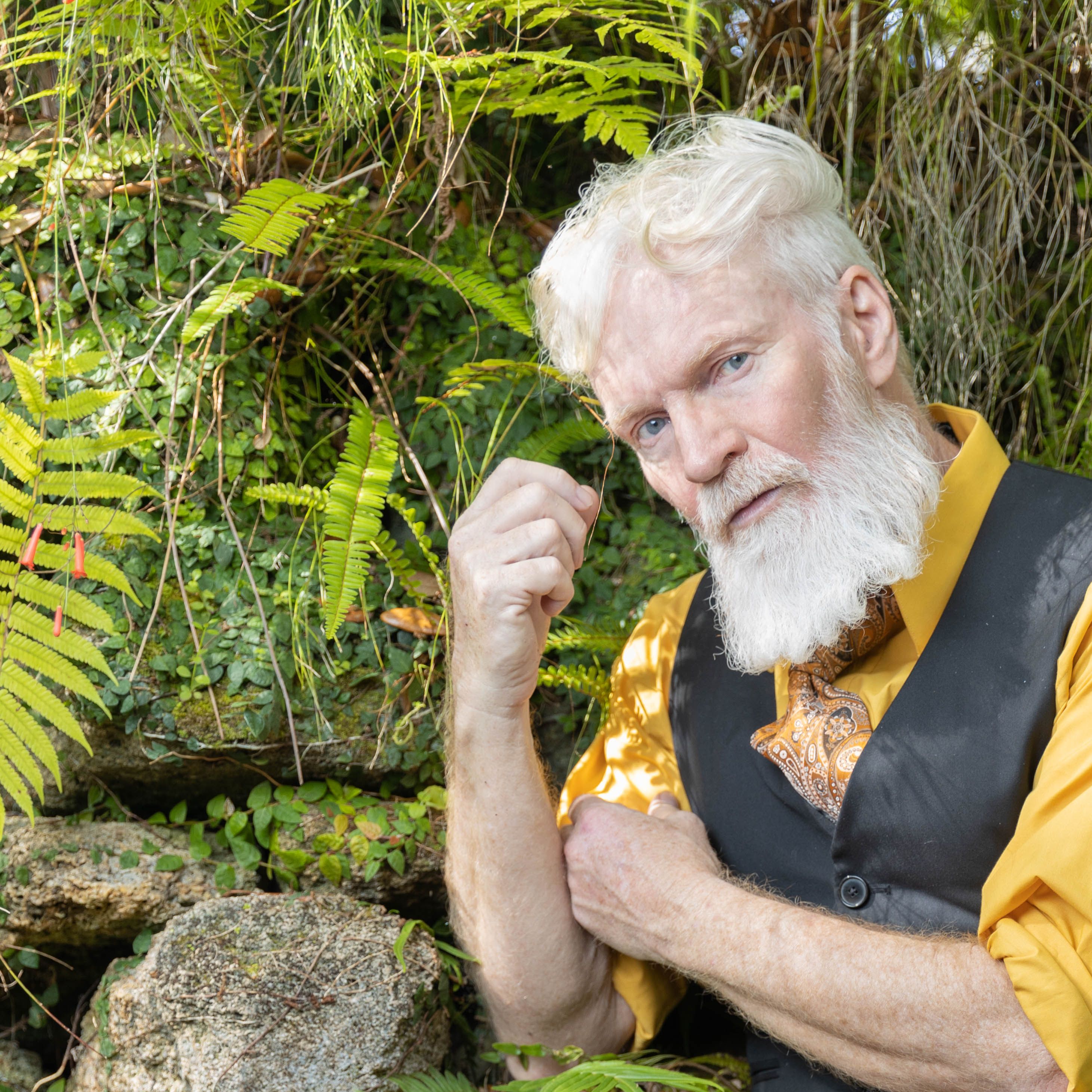 Senior man with white beard and stylish attire posing in a lush green natural setting.