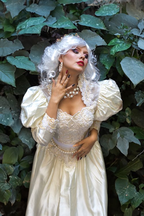 A woman in a white dress and pearls posing in front of a green plant