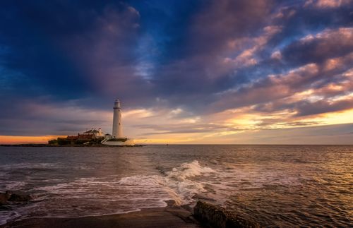 Free A beautiful sunset view of St Mary's Lighthouse surrounded by a dramatic sky and sea waves. Stock Photo