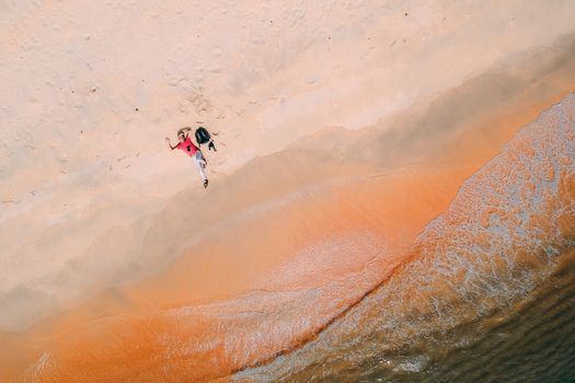 A person in a red shirt lies on a sandy beach by the ocean, photographed from above on a sunny day.