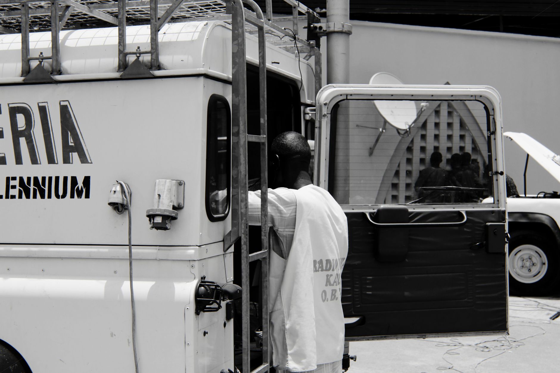 Black and white image of a man working on a vehicle in an urban Nigerian setting.