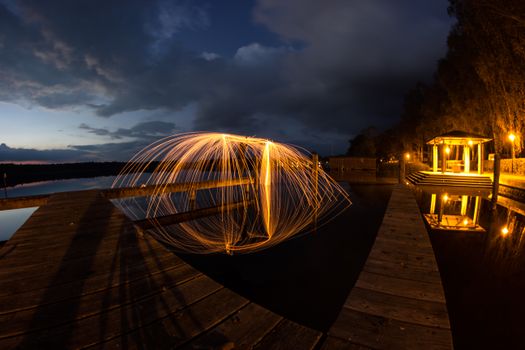 Stunning long exposure of fiery light art over a tranquil lakeside pier at dusk.