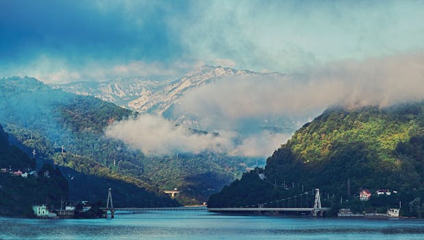 云雾缭绕的山间，一座桥梁横跨在宁静的海水上，天空多云