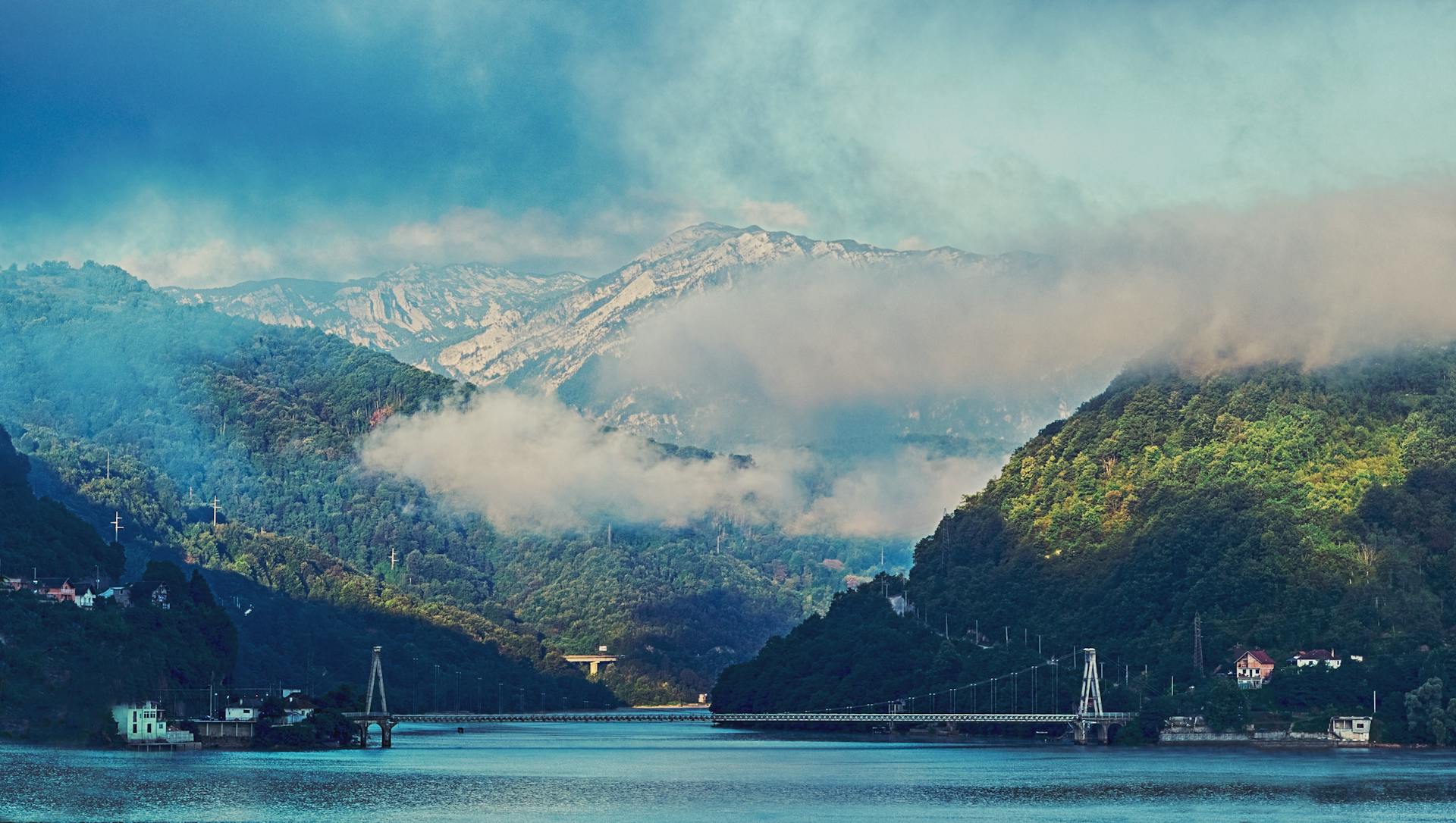 A beautiful view of a misty mountain landscape featuring a bridge and serene waters under a cloudy sky.