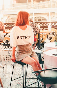 一位穿着休闲装的女子坐在酒吧凳上，享受着咖啡馆的氛围，她的衬衫上印着粗体文字
