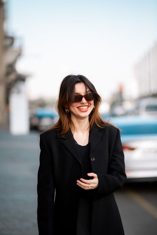 Free Stylish brunette woman smiling while walking on city street wearing sunglasses and black coat. Stock Photo