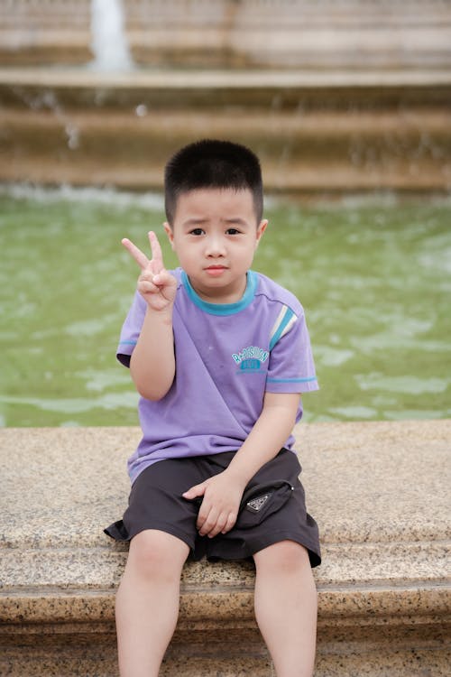 A young boy sitting on a stone ledge with his hand up