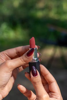 A close-up photo of hands applying red lipstick in a natural outdoor setting.