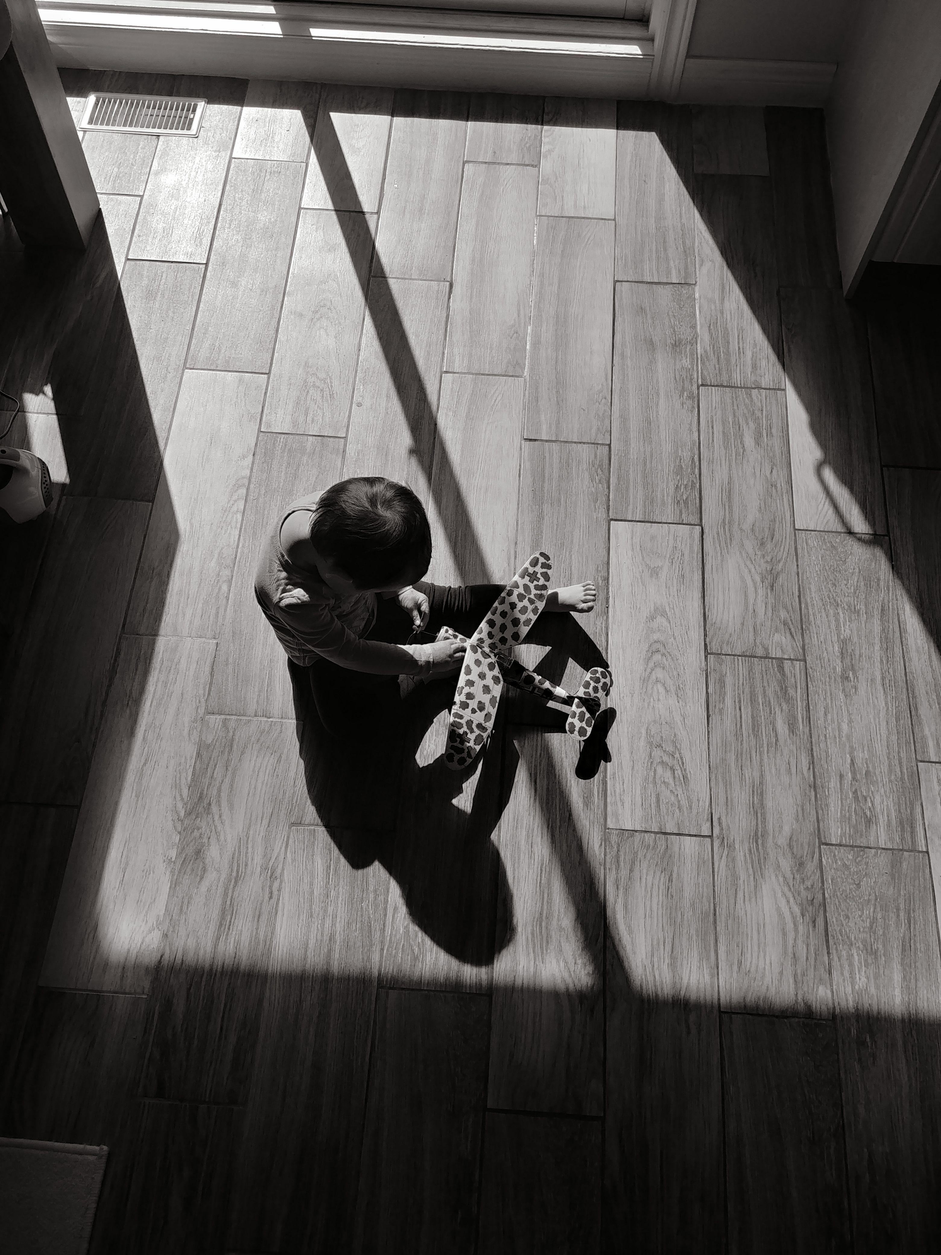 A child sits on parquet floor in sunlight, holding a toy plane, evoking nostalgia and warmth.