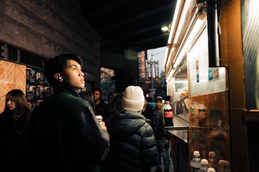 People gather at an urban street food stall under evening city lights.