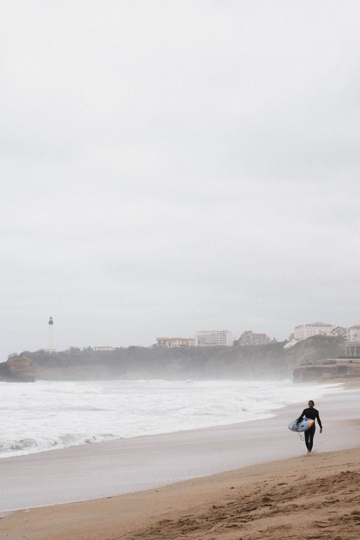 A surfer walking the Grande Plage, Biarritz lighthouse behind