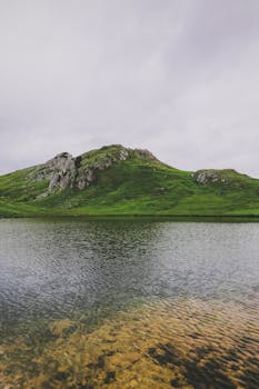 A tranquil lake with clear water reflecting a green hill in Provincia di Belluno, Italy.
