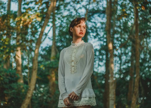 一位身穿复古连衣裙的女人站在阳光明媚的树林中，水中倒映着她的白衣身影