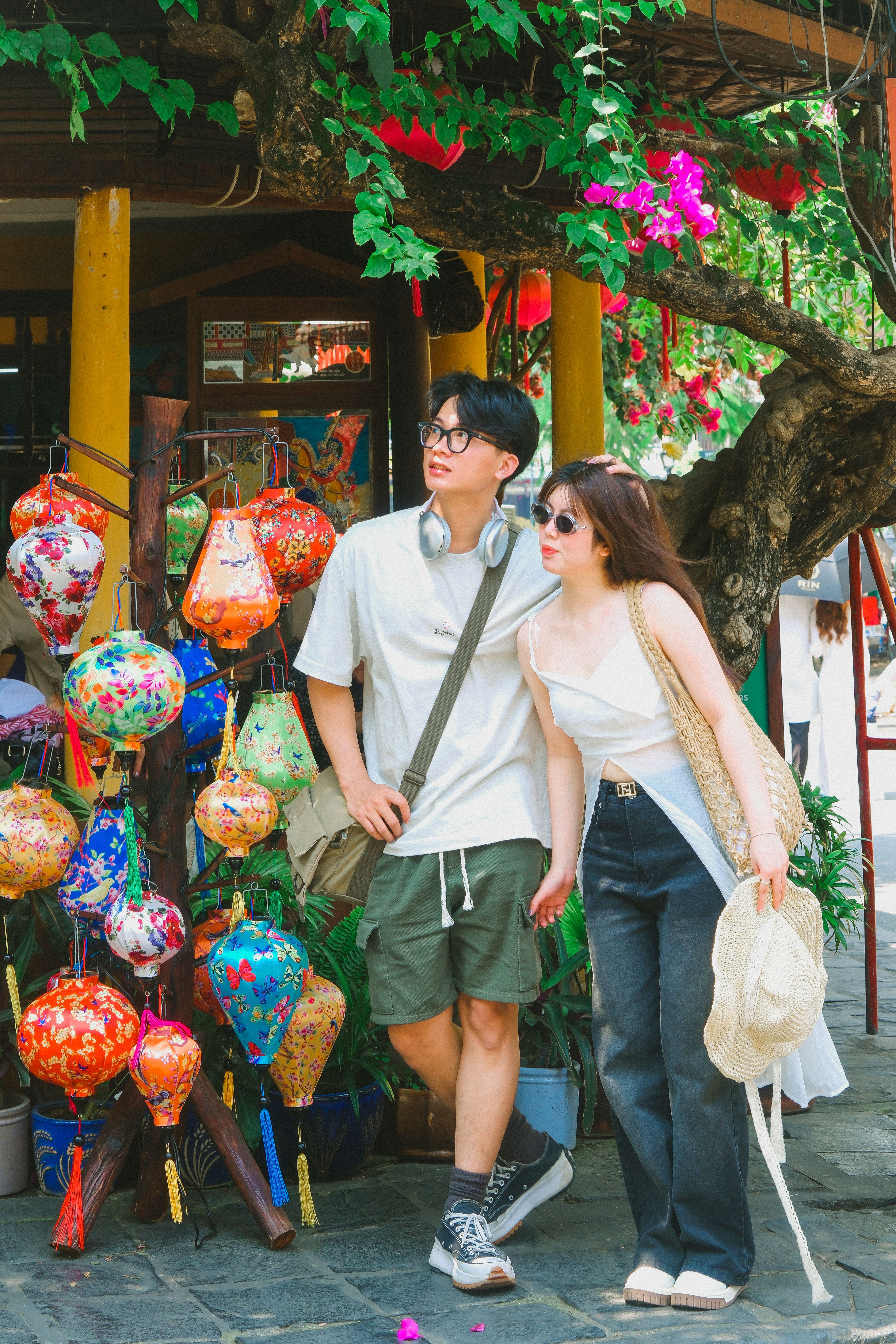 Young Asian couple exploring a colorful street market with hanging lanterns on a sunny day.
