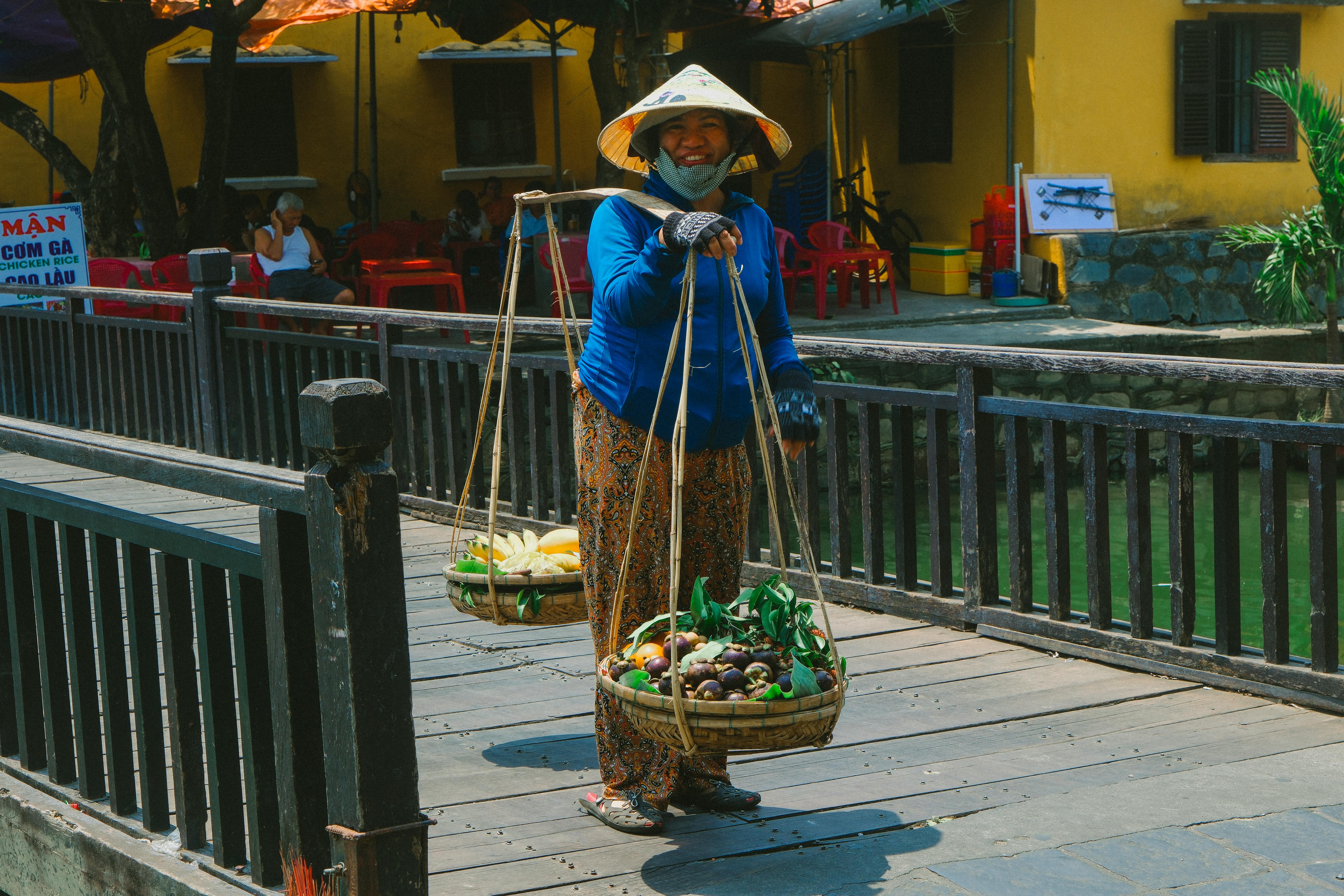 A smiling Vietnamese woman wearing a conical hat carries baskets across a footbridge on a sunny day.