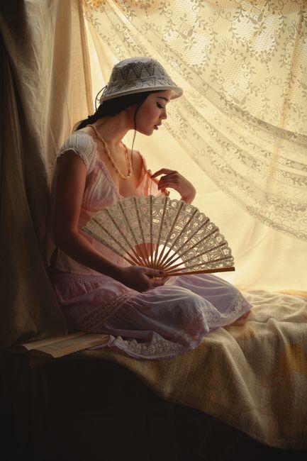 Elegant woman in vintage dress holding a fan, sitting by a window with soft light.
