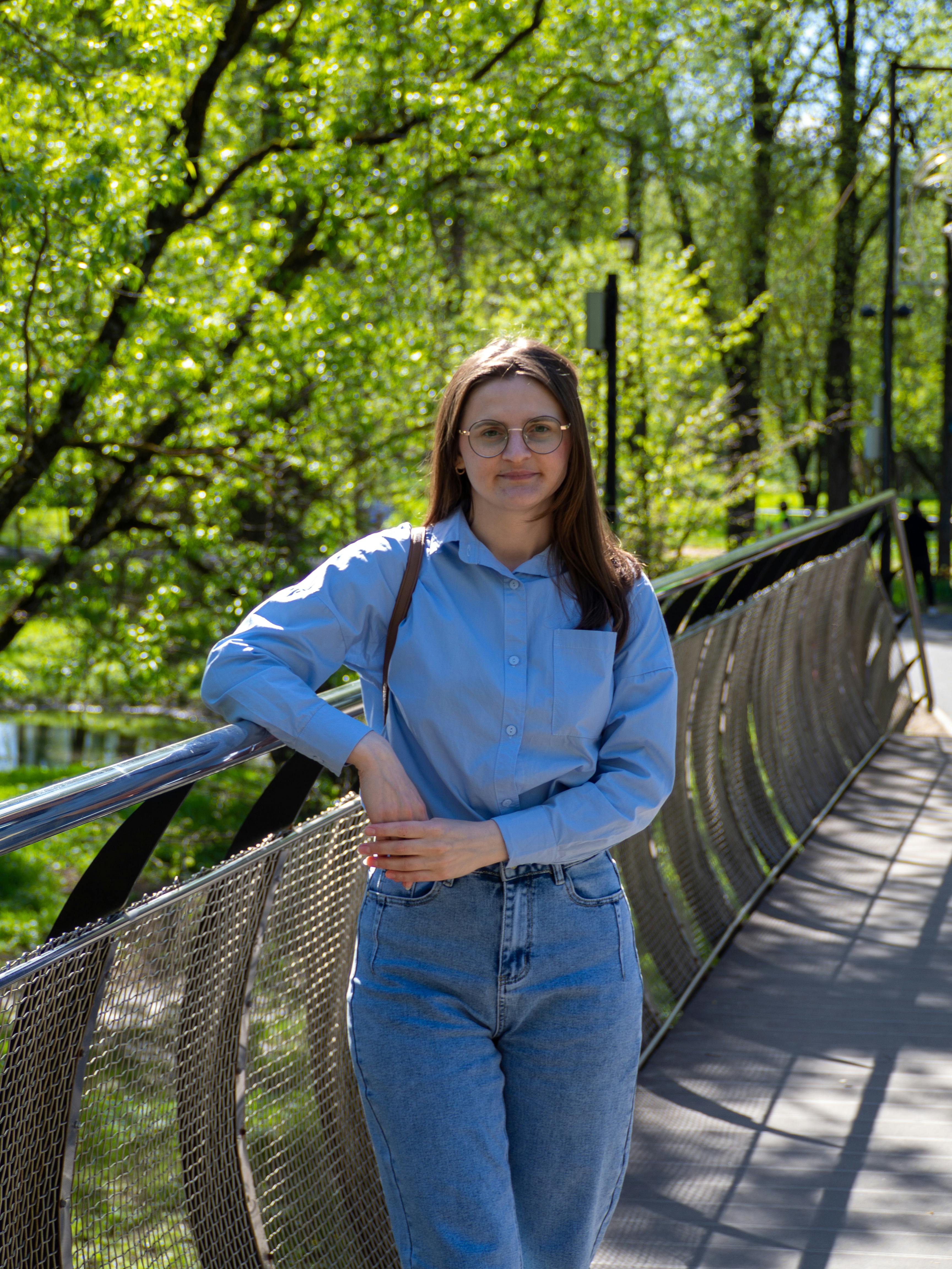 A young woman wearing jeans and a blue shirt, smiling on a park bridge during a sunny day.