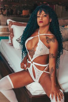 Woman in white lingerie with butterfly tattoos sitting on a bed, exuding sensuality.