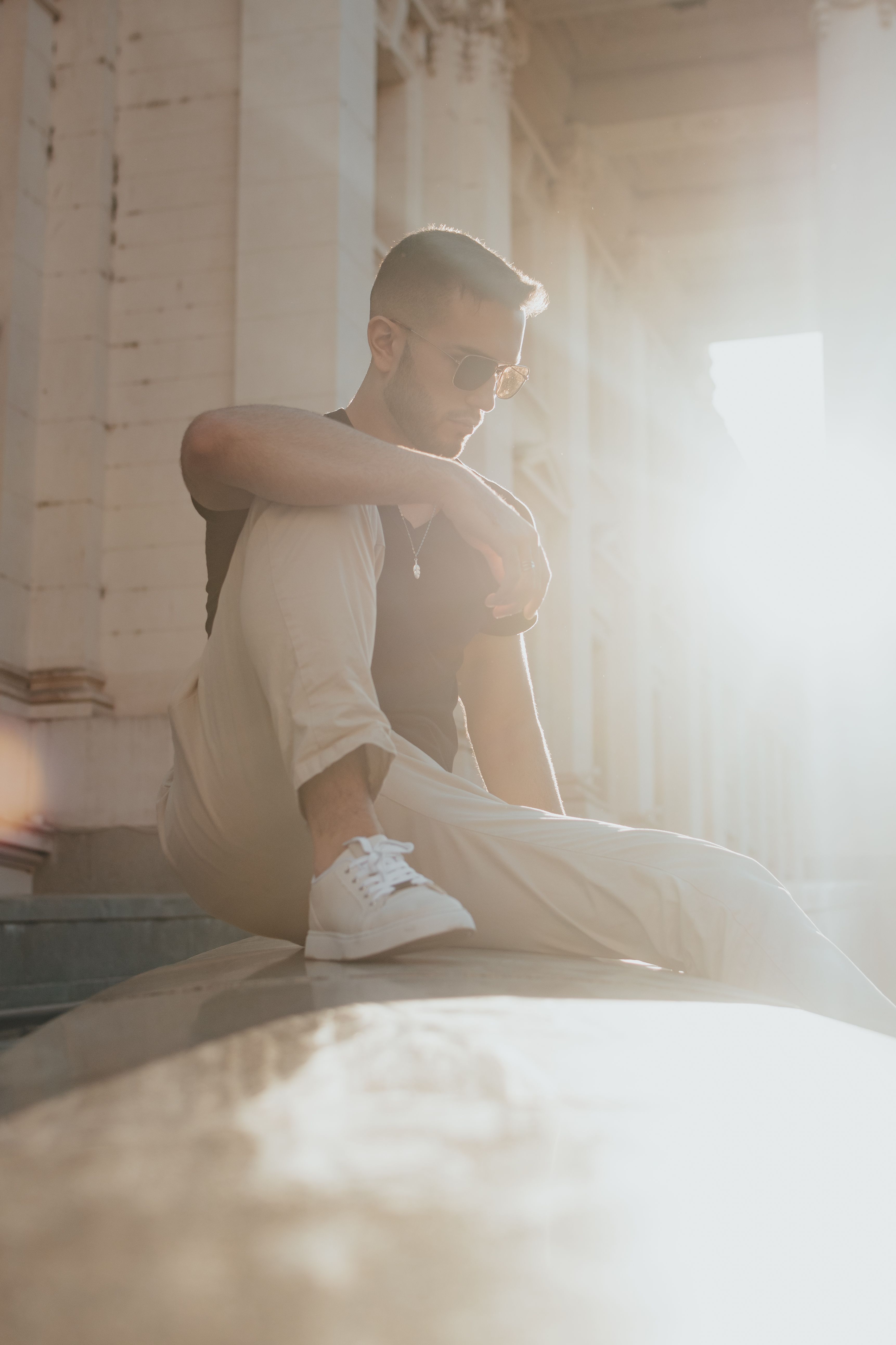 Stylish man wearing sunglasses sitting on a stone wall, soaking up sunny vibes.