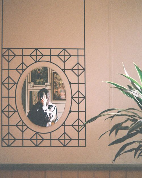 Free A woman's reflection captured in an ornate mirror alongside wall art and plant. Stock Photo
