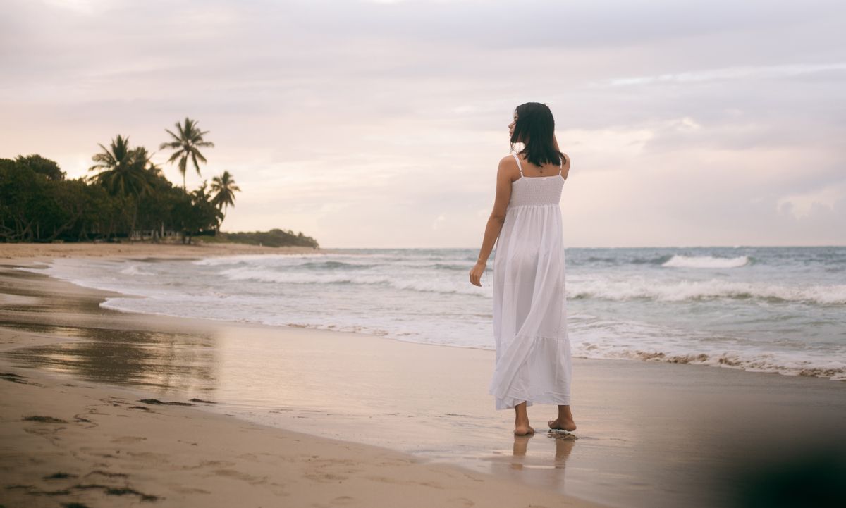 Young woman in white dress walks along Puerto Plata beach at sunset.