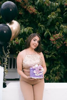 Woman in stylish outfit holding a purple cake, posed with black and gold balloons outdoors.