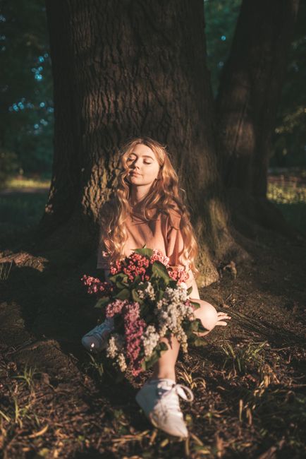 A peaceful woman sits under a tree, surrounded by flowers in an outdoor forest setting.