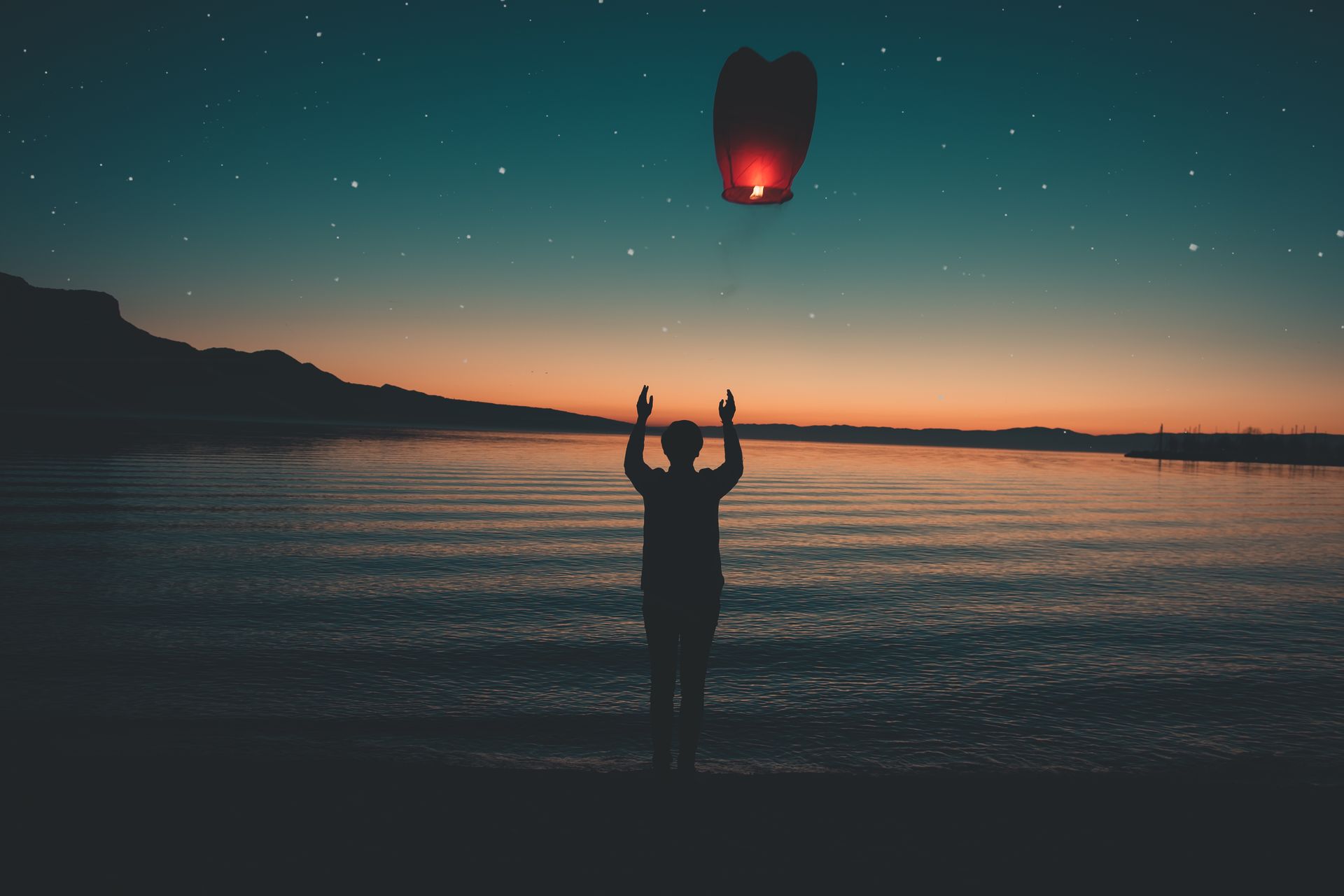 Silhouette releasing a floating lantern by a peaceful lake at sunset, creating a serene and magical atmosphere.