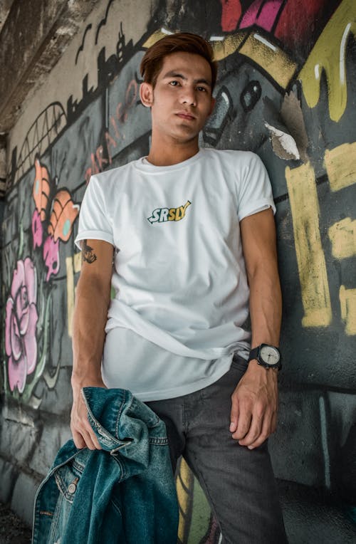 Foto De Um Homem De Camiseta Branca E Jeans Cinza, Apoiado Na Parede De Grafite, Segurando Uma Jaqueta Jeans