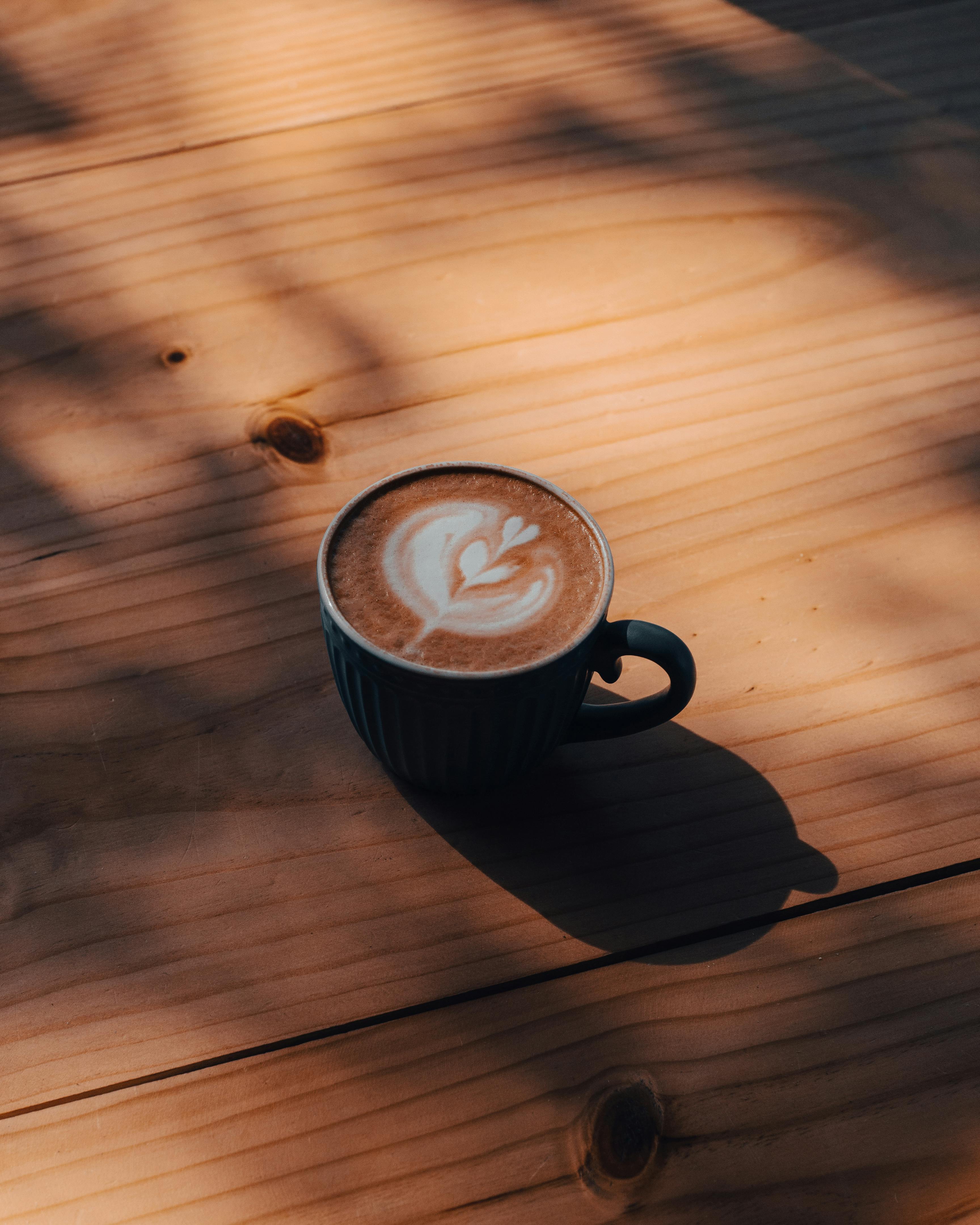 A close-up of a coffee cup with latte art on a sunlit wooden table casting soft shadows.
