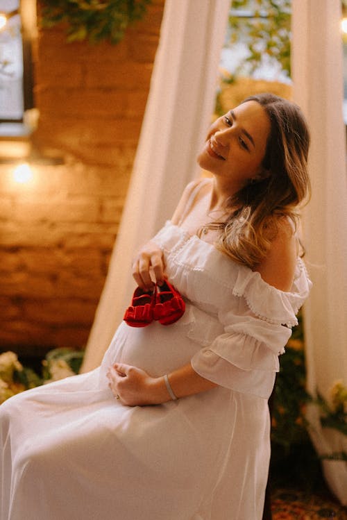A pregnant woman in a white dress is holding a red shoe
