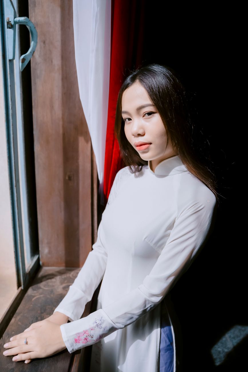 Elegant young Vietnamese woman in ao dai, posing by a sunlit window, showcasing traditional beauty and culture.