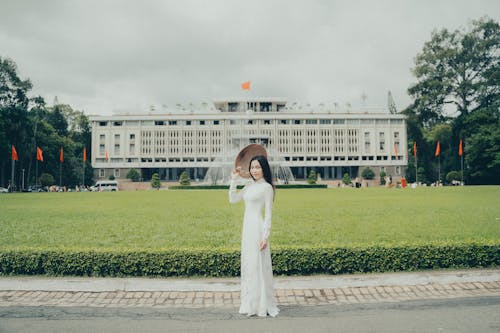 A woman in a white dress stands in front of a large building