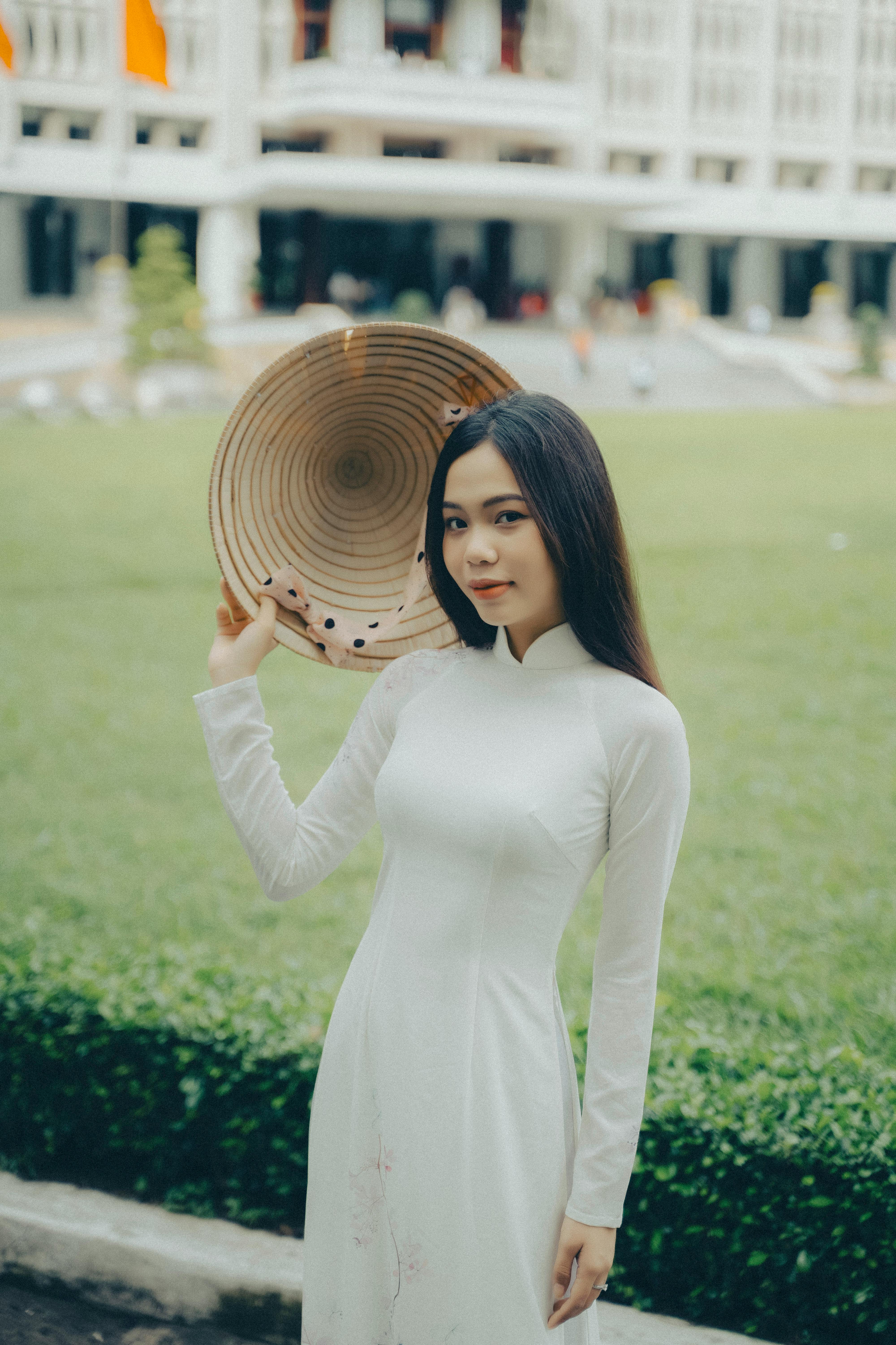 Portrait of a Vietnamese woman in a white ao dai holding a conical hat outdoors.