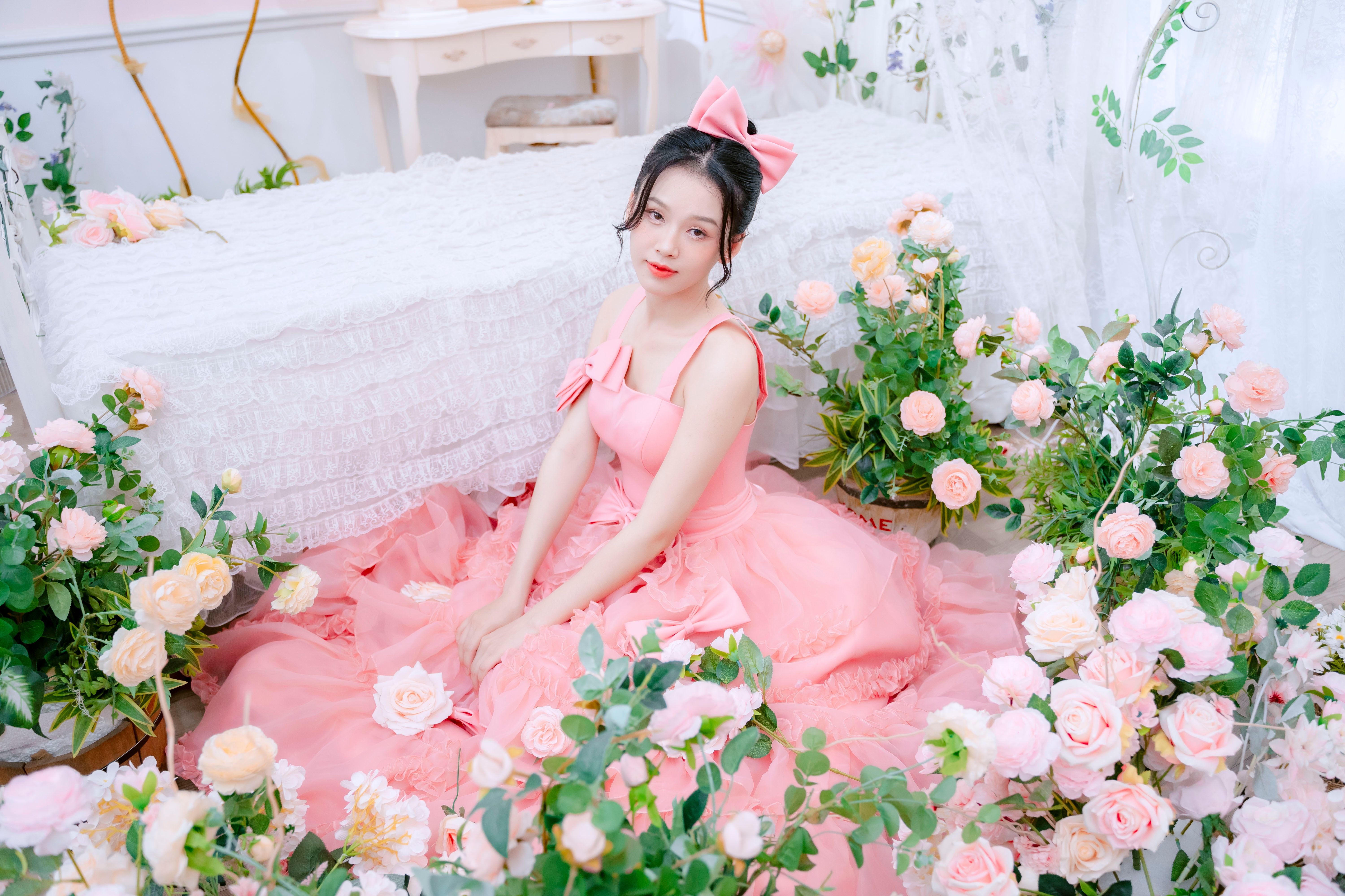 A young woman in a pink ball gown surrounded by pink roses and lush greenery.