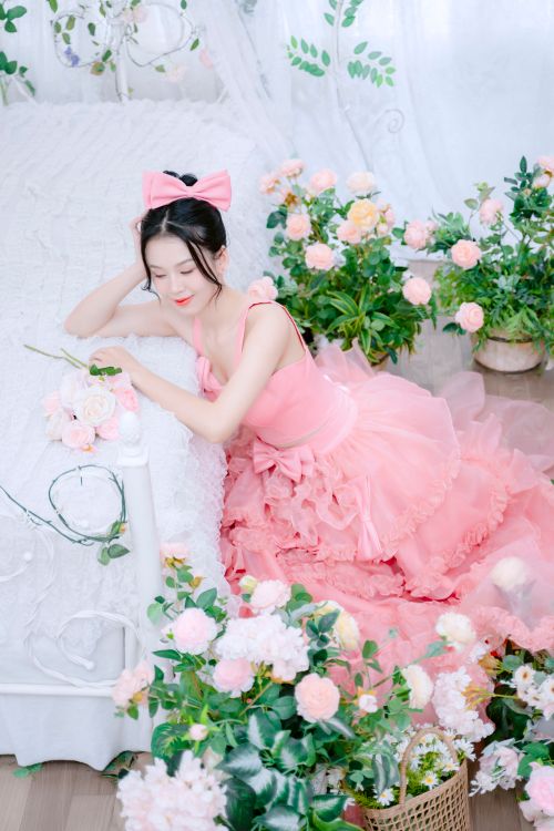 Free Elegant woman in a pink dress sitting on a bed surrounded by pink roses, smiling serenely. Stock Photo