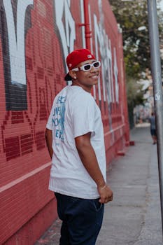 Smiling young adult in streetwear poses against graffiti wall on city sidewalk.