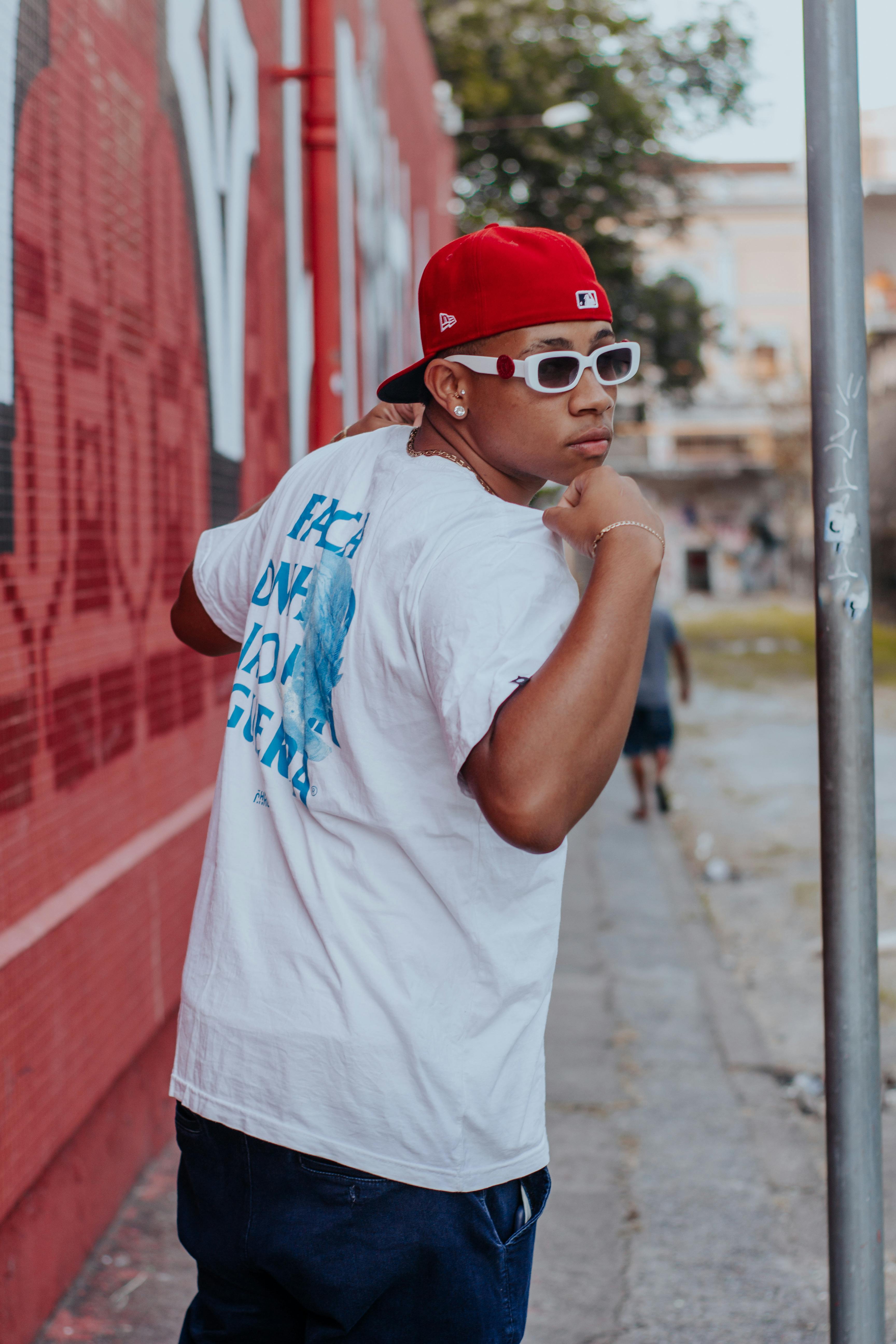 Man in casual outfit and cap poses near a graffiti wall in an urban setting.