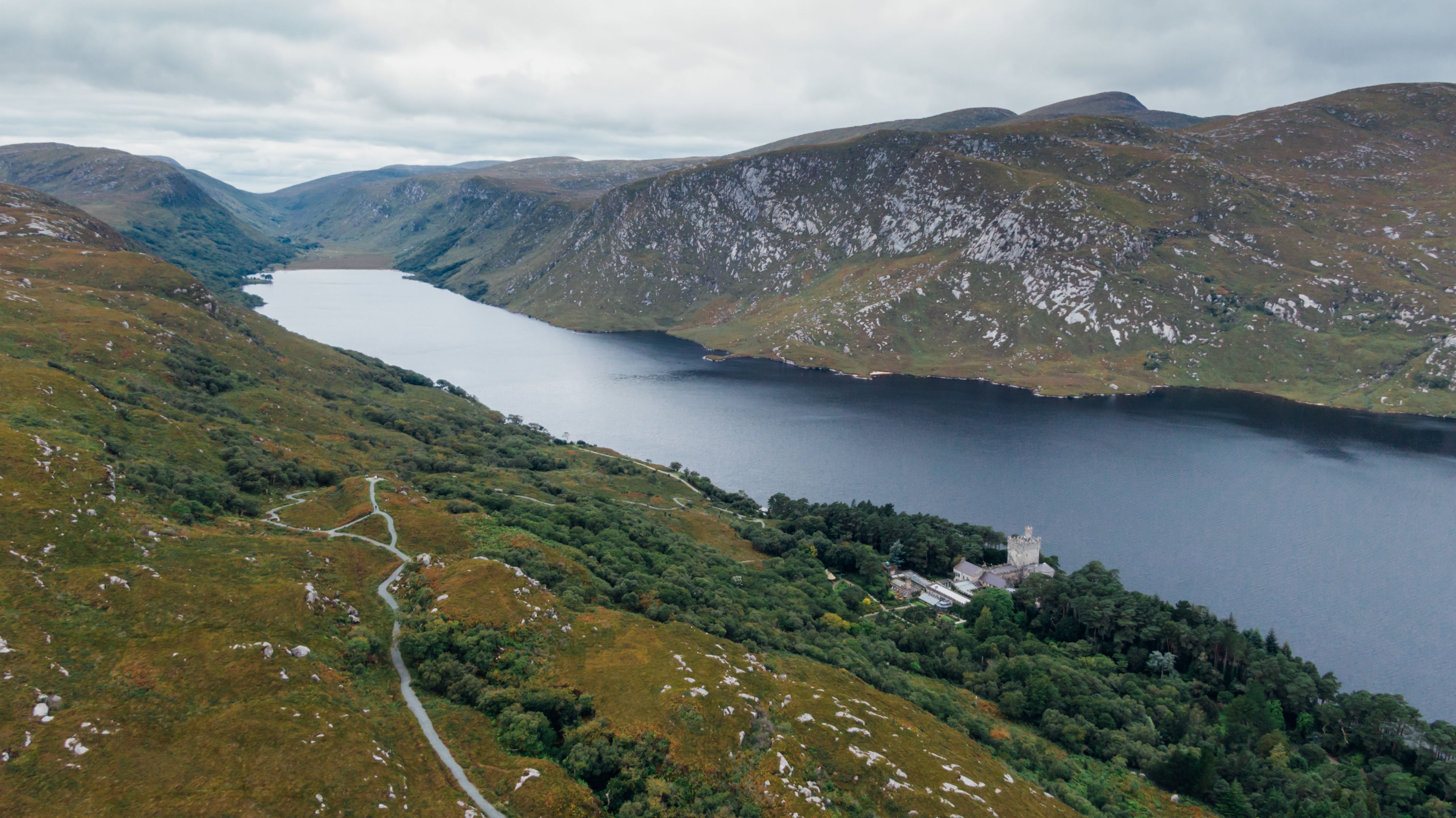 Scenic Aerial View of Donegal, Ireland’s Lakes and Hills Aerial view of the rugged green hills and serene lake of Donegal, Ireland, with a winding road cutting through the landscape and a historic stone building nestled among dense trees near the shoreline. The lake stretches into the distance between dramatic, rocky hills under a cloudy sky. Photo courtesy of Kelly via Pexels.