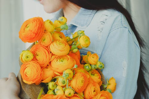 Side view of crop anonymous young female florist with long dark hair holding bunch of fresh orange flowers in hands