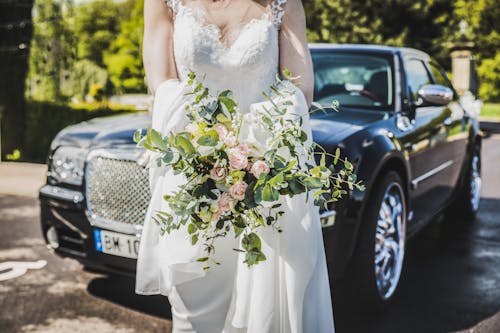 黒い車の前で花束を運ぶ白いウェディングドレスの女性