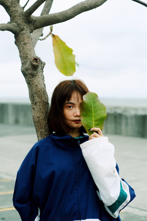 Photo of Woman in Blue and White Jacket Leaning on Tree While Holding a Large Green Leaf in Front Her Eye