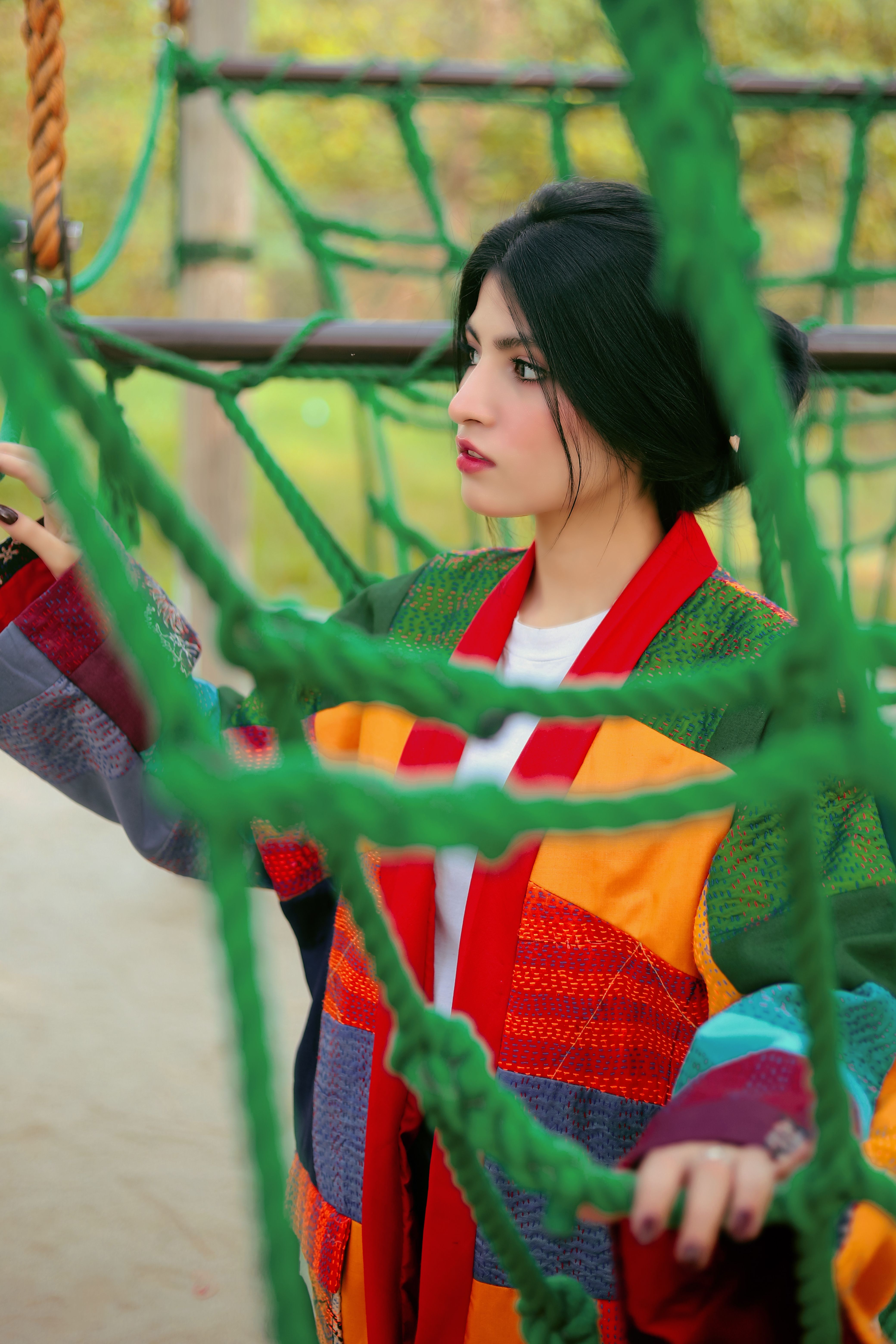 A young woman in a vibrant coat stands on a playground, framed by a green rope net.
